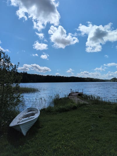This image presents a natural lakeside landscape in Hartola, Finland, under a partly cloudy blue sky. The scene is illuminated by bright daylight, suggesting late morning or early afternoon.
The upper half of the image is dominated by a clear blue sky interspersed with various white and light gray cumulus clouds. A vast, dark blue lake occupies the midground, its surface showing numerous ripples from a light breeze. The distant shoreline across the lake is characterized by a dense, dark green coniferous forest that stretches horizontally from left to right.
In the foreground, a grassy bank slopes gently towards the water. Sparse reeds and tall grasses line the immediate water's edge, partially submerged. Small yellow wildflowers are visible scattered within the grass on the right side of the foreground. A dark green, leafy bush or small tree is positioned on the far left.
A light-colored, possibly white or pale gray, small rowboat or dinghy is pulled onto the grass in the lower-left quadrant, oriented diagonally towards the lake. Oars or similar wooden items are visible within its hull. A wooden dock, constructed from planks, extends into the lake from the grassy bank on the right side of the foreground. A simple, light-colored wooden chair is placed at the far end of this dock, facing the expanse of the lake. No individuals are visible anywhere in the depicted scene.
