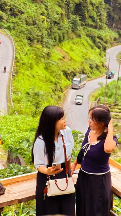 Two women stand on a wooden platform overlooking a winding road in what appears to be a mountainous or hilly area. Lush green vegetation covers the landscape surrounding the road.
The woman on the left wears a light-colored, collared shirt, a dark skirt, and glasses. She holds a smartphone in her hand and has a white purse with brown accents over her shoulder. The woman on the right is dressed in a dark blue top with a white-trimmed neckline and a dark skirt. She is touching her hair and also wears glasses.
The road below features several vehicles, including trucks and a motorcycle. The scene suggests an elevated viewpoint or a scenic overlook, likely in a rural or semi-rural area given the abundance of greenery.
Based on the prompt, the location of this scene is in the city of Phố Bảng Commune, Vietnam.