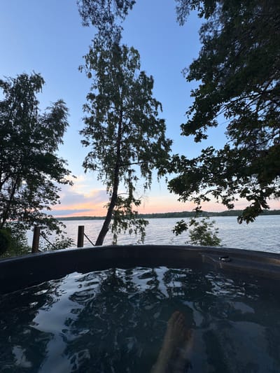 The image captures a serene outdoor scene at dusk, likely in Priedesmuiža, Latvia. In the foreground, the dark, rippling surface of water fills a portion of a dark-colored hot tub. A bare foot is partially submerged in the water on the right side of the frame. Beyond the hot tub, a wooden railing with vertical posts separates the viewer from a body of water, which appears to be a lake. The lake stretches to the horizon, where a soft, muted sunset paints the sky with hues of pink, orange, and blue. Silhouettes of trees frame the upper portion of the image, their branches reaching into the twilight sky. The overall atmosphere is tranquil and inviting, suggesting a moment of relaxation in nature.