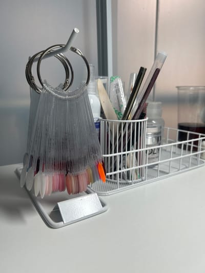The image shows a white wire basket mounted on a white wall, likely inside a building. The basket contains various nail care items, including brushes, nail files, and bottles with labels. In front of the basket, attached to a metal rod, are three metal rings holding numerous clear plastic nail color sample sticks, displaying various nail polish colors. The colors range from neutral tones to pink and white. The scene appears to be in a salon or personal care setting. There is no evidence suggesting the location is specifically in Аксукент, Kazakhstan.