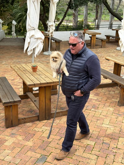 A man is standing outdoors on a brick-paved area in Theewaterskloof Nu, South Africa. He is wearing sunglasses, a dark blue sweater, a quilted vest, dark blue jeans, and brown shoes. He is holding a fluffy, light brown dog with a gray leash. The man has his right hand in his pocket. Behind him are wooden picnic tables and benches, some with folded white umbrellas. In the background, trees and greenery are visible.