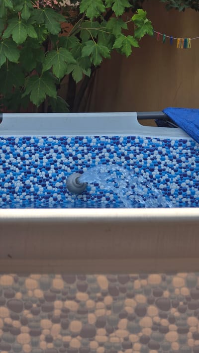 In Kutná Hora, Czechia, a close-up shot captures a water jet spraying from a grey nozzle into an above-ground swimming pool. The pool liner is decorated with a mosaic pattern of blue and white circles. In the background, green leaves from a tree are visible, and a clothesline with colorful clothespins hangs on a tan wall. A blue tarp is draped over part of the pool's frame. The foreground shows the edge of another surface, also with a similar mosaic pattern.