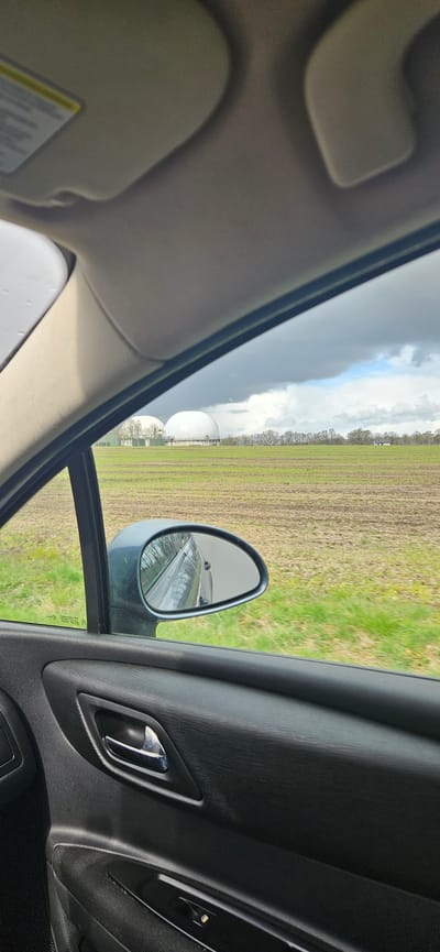 The image shows a view from inside a car, likely a sedan, looking out the passenger-side window. The car's interior includes a tan-colored sun visor, a dark gray door panel with a silver door handle, and a side mirror. Outside, the scene appears to be a rural or agricultural area in Delbrück, Germany. In the distance, a large cultivated field is visible, transitioning from green to brown, with rows indicating possible planting or tilling. A line of trees borders the field. A series of spherical white tanks or containers are partially visible on the left side of the frame near the field. The sky is overcast with a mix of dark storm clouds and patches of white clouds. The mirror reflection shows a road and presumably the car traveling on it.