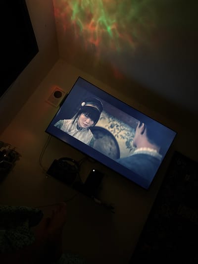 The image depicts a dimly lit room with a television screen displaying a scene from a show. The TV is mounted on a wall, and on the wall above it, there are colorful light projections, appearing as swirling green and red patterns. To the left of the TV, a white square object is mounted on the wall, possibly a speaker or sensor. Below the TV, a black gaming steering wheel and pedals are visible, along with a black electronic device. In the foreground, a person's foot, wearing a patterned blanket, is visible in the lower left corner, suggesting someone is relaxing and watching the television. The overall environment appears to be a personal living space, possibly a bedroom or living room, set up for entertainment. The image does not contain any identifiable landmarks or information to place it specifically in Santa Marta, Colombia.
