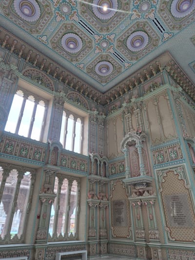 This image depicts the interior of the BAPS Swaminarayan Mandir in Robbinsville Township, New Jersey, United States of America. The scene is an ornate hall with intricately decorated walls and ceiling. The walls are adorned with detailed carvings, patterned textures, and colorful motifs, featuring elements of pale pink, blue, and cream. There are several large, arched windows with translucent panes, allowing natural light to filter into the space. The ceiling is also elaborately decorated with geometric patterns and floral designs in similar pastel colors. Various architectural features, such as columns, arches, and niches, are visible throughout the hall, contributing to the overall elaborate and decorative aesthetic. Inscriptions in what appears to be Sanskrit or Hindi are present on some of the wall panels. There are no people visible in the image.