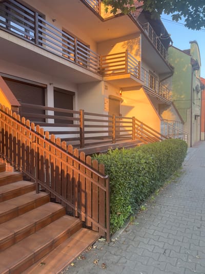 This media file shows an exterior view of a multi-story building with a light green facade on the upper floors and a cream-colored facade on the lower floors. There are multiple outdoor staircases with wooden railings and treads, leading up to different levels of the building. The stairs and railings are illuminated by warm, golden sunlight, suggesting it is either early morning or late afternoon. A dense, green hedge runs along the side of one of the staircases. The ground adjacent to the hedge and stairs is paved with grey interlocking stones. A portion of a mature tree with green leaves is visible in the upper left corner of the frame. The building appears to be residential. There is no visible text in the image. The scene is peaceful and bathed in the soft light of a setting or rising sun. The location is Skawina, Poland.