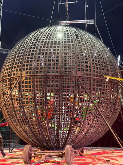 The image shows a metal globe-shaped structure, likely a "Globe of Death" performance cage, illuminated by bright spotlights. Inside the globe, there are indications of people wearing colorful clothing and possibly riding motorcycles. The globe is made of interconnected metal bars creating a grid pattern. The backdrop includes a dark night sky on one side and a red and white striped tent-like structure on the other. A tire is mounted next to the tent structure. The location is identified as Tejalpa, Mexico.