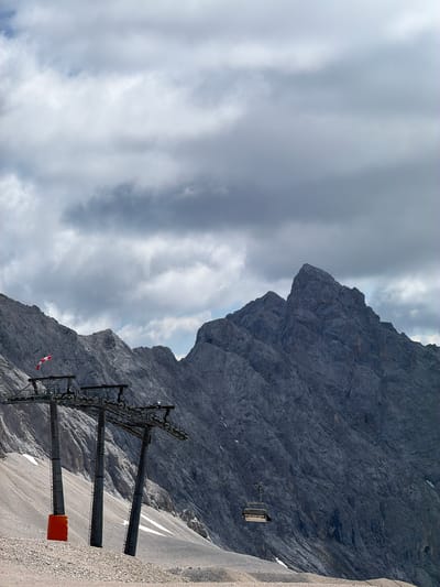 This is an image of a ski lift against a dramatic mountain backdrop under a cloudy sky. The ski lift has three tall, grey pylons supporting the cables and a single gondola is visible hanging from one of the cables in the middle of the frame. In the foreground, a red marker is visible at the base of the leftmost pylon. A red and white striped flag, possibly a windsock, is attached to one of the pylons. The mountains themselves are rugged and rocky, with patches of snow. The sky is filled with dramatic, grey clouds, suggesting overcast or stormy weather. This scene is located in the city of Wildermieming, Austria.