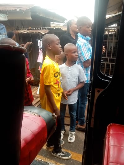 The image, taken in Umuokpu, Nigeria, shows a view from inside a vehicle, possibly a taxi or bus. Three young boys stand outside, looking in the same direction. The boy closest to the camera wears a yellow t-shirt, another wears a gray button-down shirt and the third is wearing a blue plaid shirt. An African woman is standing next to the children and has her hair pulled back. The surroundings are urban, with a building constructed of corrugated metal and wooden beams with a mix of clothing hanging and miscellaneous items scattered around. A woman with red clothing on and someone holding a rubber band are visible behind the boy in yellow. Inside the vehicle, a worn, red-and-black seat is visible.