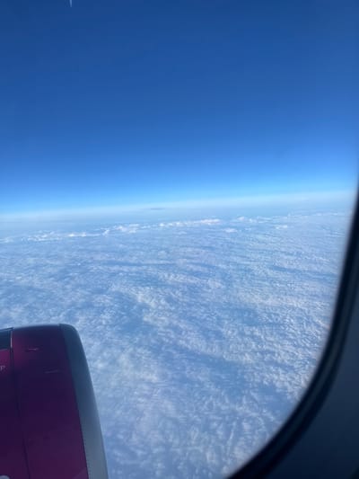 The image presents an aerial view captured from an aircraft window, looking out over an extensive layer of white, textured clouds. The sky above is a clear, deep blue, transitioning to a lighter, hazy blue near the horizon. In the lower-left quadrant, a portion of an aircraft engine nacelle is visible; its primary color is a distinct purple/magenta, with a segment of its outer casing appearing grey or silver. The curved, dark grey frame of the aircraft window is visible along the right edge, partially framing the scene. No people, specific actions, or interactions are depicted. The environment is an upper atmosphere perspective, completely above ground features. Due to the high altitude and complete cloud cover, no specific geographic landmarks, including those of Caselle Torinese, Italy, are discernible within the image.