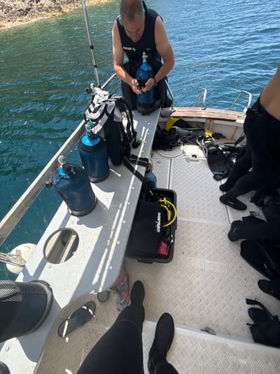 A man in a black wetsuit with "BEUCHAT" visible on the shoulder is on a boat, holding a blue scuba tank. Two other blue scuba tanks are standing upright on the boat's deck. Divers' fins, a black equipment bag labeled "SUPRAS" and yellow hoses are also visible on the deck. The boat is in the water with a rocky shoreline in the background. The scene suggests preparations for a diving excursion in the waters near Potos, Greece.