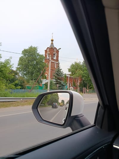 The image is taken from inside a moving vehicle, looking out the side window and into the side mirror. In the foreground, the edge of the car's interior is visible, along with the black frame of the side mirror and the white casing of the mirror assembly. The side mirror reflects a view of the road behind the vehicle, showing a gray asphalt road with a white dividing line, a few cars, and trees. The sky in the reflection appears overcast and pale.

Outside the car, and to the left of the side mirror, a red brick church with a golden dome and cross is visible. The church has a bell tower with arched openings and decorative white elements. Trees with green foliage surround the church, and a blue wooden fence runs along the side of the road in front of it. A street lamp is also visible near the church. The road outside the car is gray asphalt with a white line, and there are trees and vegetation on the right side of the road.

The image appears to be taken in a rural or suburban setting in Russia, possibly in or near the city of Речицы, based on the style of the church and the surrounding environment. No people are clearly visible in the main part of the image or in the reflection. The action depicted is passive, showing a scene observed from a moving vehicle.