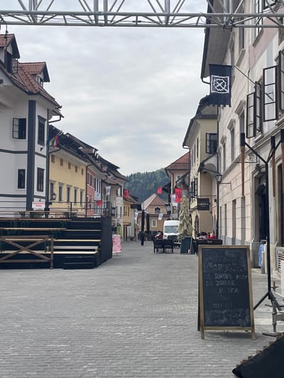 The image shows a street in Skofja Loka, Slovenia, possibly preparing for a festival or event. The street is lined with buildings of varying colors and architectural styles. Banners and flags are hung between buildings, including one with a distinctive black and white emblem. There are structures like a stage or platform constructed on the right side of the street. A blackboard displaying text in Slovene, possibly a menu or event schedule, is also visible on the right. The street is paved with cobblestones, and some pedestrians are present in the background. Metal scaffolding is visible on the left. A forested hill is visible in the distance.