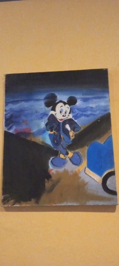 The image is a vertically oriented photograph of a rectangular painting, reportedly located in the city of Aral, Kyrgyzstan. The painting depicts a stylized, full-body rendition of the cartoon character Mickey Mouse positioned centrally. Mickey Mouse is shown facing slightly towards the viewer's right, wearing a dark blue jacket with a white collar, blue lower garments, and traditional yellow shoes. His right hand, covered by a white glove, holds a slender white object. His facial expression is a slight smile.
The painting's background features a dark blue sky above a turbulent body of water, rendered with prominent light blue and white waves. The immediate foreground around Mickey Mouse consists of dark, uneven terrain or abstract shapes, with a significant dark mass on the left. To the right, a large dark blue shape, possibly a structural element or part of a vehicle, is visible, including a partial black circle at its base. Subtle patches of red, light brown, and grey coloration are present on the ground plane. The painting itself is positioned against a flat, uniform light orange-yellow surface, which serves as the background of the photograph.