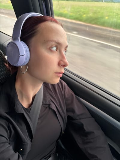This medium close-up image, oriented horizontally, captures a young individual inside a moving vehicle, likely a car or bus, in Kvarsa, Russia. The subject, who appears to be female, is positioned looking out of a side window. She has light skin, reddish-brown hair, and long eyelashes.

She is wearing light purple over-ear headphones, with the "JBL" brand clearly visible on the left earcup. A silver, textured hoop earring adorns her left ear. Her attire consists of dark clothing, possibly a black jacket or shirt, and a black seatbelt crosses diagonally over her chest. Her expression is neutral and contemplative, with her gaze directed upwards and outwards, possibly observing the passing scenery or lost in thought. Bright reflections are visible in her eyes.

Outside the window, a blurred landscape of green foliage and a road or pathway suggests movement and travel. The lighting indicates it is daytime. The overall mood of the image is quiet and introspective, capturing a moment of rest or reflection during a journey.