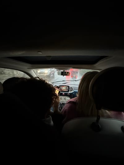 Inside a vehicle, two people are visible. The driver, with blonde hair and wearing headphones, looks towards the passenger. The passenger, with curly hair, holds a smartphone. Through the rain-streaked windshield, the faint outlines of other vehicles are discernible. The dashboard is partly visible, featuring a navigation system and various buttons. The interior of the car is dark, creating a stark contrast with the faint external light.