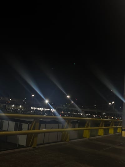 At night in Rionegro, Colombia, a parking lot illuminated by streetlights is seen from a slightly elevated position. Yellow safety railings run along the right side of the frame, partially obscuring a building with visible windows and mesh panels. Beyond the railings, rows of parked cars stretch into the distance, lit by more streetlights. The sky above is dark, with light flares around the streetlights.