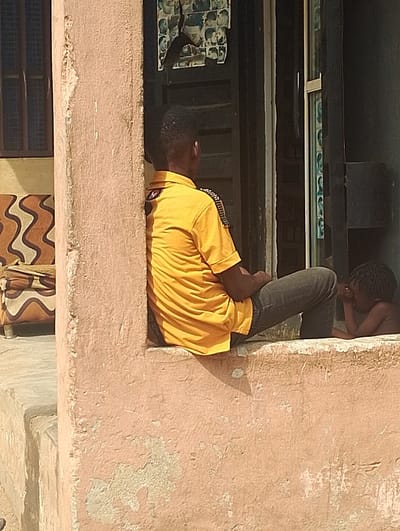 The image shows a scene in Ogharefe, Nigeria, located at 5.935 latitude and 5.67 longitude. A man is sitting on a ledge, partially obscured by a pillar, wearing a yellow t-shirt and blue jeans. His right leg is extended, and his left leg is bent with his left hand resting on it. His face is obscured by the pillar. He is looking to the right. Another person, likely a child, is sitting on the ledge behind him, partially out of frame. This person is wearing a black shirt, and is looking down and to the left, with their left hand on their lap and their right hand in their lap. Their face is partially visible, and they appear to be looking down at their hand. There is a window and doorway behind the individuals. In the window, a glass pane is partially visible, and there is a series of photographs of men's hairstyles visible on the wall outside the window. The building is made of beige concrete, with a worn, faded appearance. The background consists of a portion of a wall and a door. There is a cushion on the ground in the bottom left corner of the frame. The scene is brightly lit and there are no other visible details.
