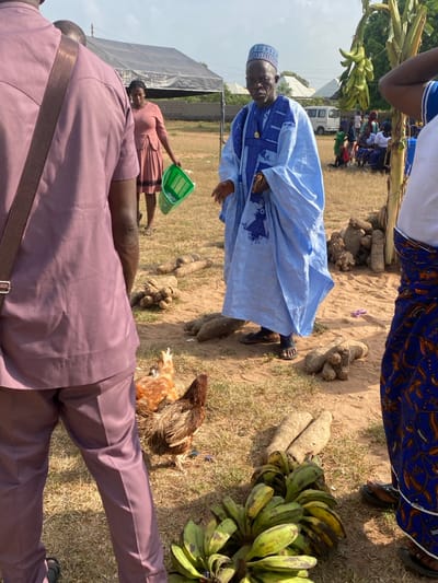 This image depicts an outdoor scene in Itobe, Nigeria, under a bright sky. In the foreground, the back of a person wearing a maroon-colored suit is visible on the left. To their right, two chickens, one reddish-brown and one dark brown, stand on dry, light brown ground. A large bunch of green and yellow plantains rests on the ground directly in front of the person in maroon. Several large, cylindrical yams are scattered across the mid-ground.
In the center of the image, an older man with a beard, wearing a light blue and dark blue patterned caftan and matching cap, stands facing slightly left, with his hands open in a gesturing posture. Behind him and to the left, a woman in a pink dress walks away from the viewer, carrying a bright green plastic basket in her left hand.
The background features a black canopy tent on the left. Further back, a white van is parked, and a group of people are seated under what appears to be another gathering point or tree. The ground throughout the visible area is dry and dusty.