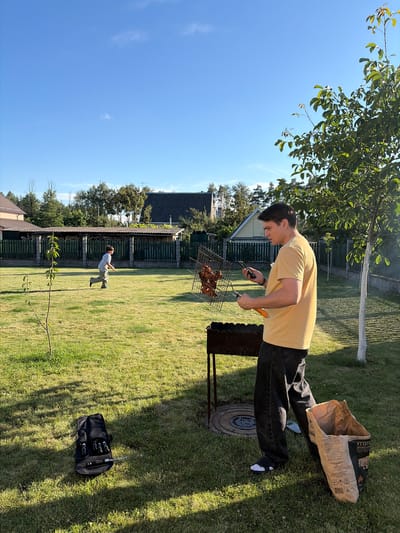 In the village of Navadvorski Sel'ski Savet, Belarus, on a sunny day, a man stands in a grassy backyard, holding a phone in his left hand and a grill rack with chicken pieces in his right. He is wearing a yellow t-shirt and dark pants. Beside him is a barbecue grill and a large bag of charcoal. In the background, a child runs across the lawn, and houses with fences are visible. A black bag containing grilling tools lies on the grass in the foreground.