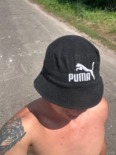 A shirtless individual wearing a black bucket hat with a white Puma logo stands outdoors on a paved surface, possibly a road or path, with greenery visible to the right. The person has a tattoo on their left arm and skin redness on their chest and shoulders. The scene is illuminated by daylight.