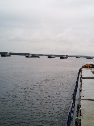 The image shows a view of the Shestovo river in Russia, on a cloudy day. Several barges are lined up on the water, with land and trees visible in the distance. The perspective is from the deck of another vessel, which is partially visible in the foreground on the right side of the image, showing the edge of its deck and some cargo containers. The water is calm, with small ripples. The sky is overcast with a thick layer of clouds, creating a muted, gray light.