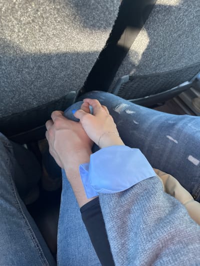 Two individuals are depicted holding hands inside what appears to be a vehicle in La Estación, Spain. The image is taken from an overhead perspective, looking down at their laps. The subject on the left wears plain blue denim jeans, with their right hand visible. The subject on the right wears ripped blue denim jeans, with their left hand, wrist, and forearm visible. This individual's left hand has blue nail polish on the fingernails and a silver chain bracelet on the wrist. Their arm is clad in a light blue shirt cuff emerging from a grey knitted sweater. Their hands are clasped together, fingers intertwined. In the background, a grey fabric seat back is visible, partially illuminated by sunlight, suggesting a window nearby. Darker areas beneath the seat back suggest the vehicle's floor or footwell.