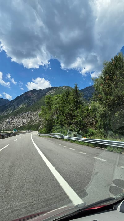 This is a sideways, eye-level shot taken from inside a vehicle, looking out at a mountain road. The foreground shows the asphalt road with a thick white line marking the lane. A metal guardrail with white stripes runs along the side of the road, separating it from a steep, rocky mountainside covered in lush green trees. The sky is a vibrant blue, dotted with fluffy white clouds and larger, darker grey clouds. The sun appears to be shining, suggesting it's daytime. The scene evokes a sense of travel and scenic beauty.
The image is captured from a moving vehicle, likely a bus or RV, as indicated by the reflections in the window. There are no people visible in the frame. The road appears to be a two-lane highway, curving gently into the distance. In the far distance, several cars can be seen traveling in the same direction as the viewer. The mountains are imposing and rugged, with some bare rock faces interspersed with dense foliage. The overall impression is one of a picturesque journey through a mountainous region.
There is a small orange marker on the side of the road near the guardrail. A circular sticker on the vehicle's window reflects in the image, showing the word "DOCKERS" in black text. The location context indicates this scene is likely near Pontebba, Italy. The weather appears to be partly cloudy and pleasant, suitable for driving and enjoying the scenery.
