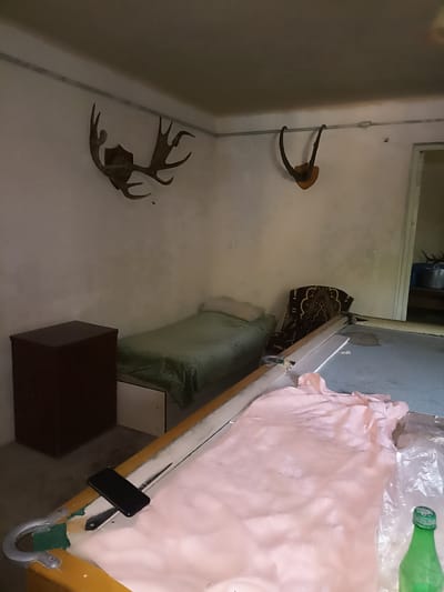In a room in Aygezard, Armenia, a single bed covered with a green blanket is positioned next to a dark brown cabinet. Above the bed, a large set of antlers is mounted on the wall, and to the right, another, smaller set of antlers is also displayed. A pool table occupies a significant portion of the room, with a pink sheet draped over its surface. A black smartphone rests on the edge of the pool table. A green bottle with a white cap is visible on the right side of the image, partially obscured by plastic sheeting. The room appears to be dimly lit, with pale walls and a neutral-colored ceiling. The overall impression is of a functional, possibly rustic, living space with decorative elements related to hunting or nature.