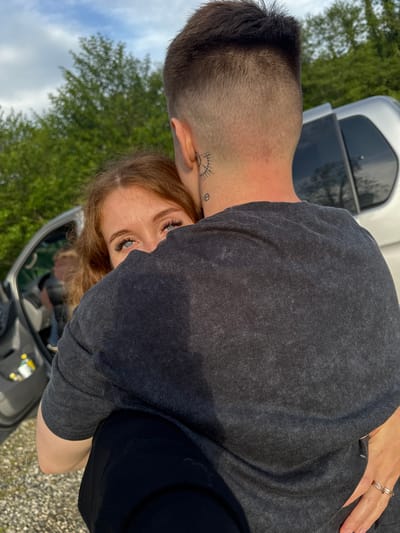 A medium shot shows a man and a woman. The woman has long reddish-brown hair and blue eyes. The man has very short, dark hair and a tattoo behind his ear. He wears a gray t-shirt. Both are standing outside of a car. The setting appears to be an outdoor area with trees in the background. No evidence suggests this is Abzhanda, Abkhazia.