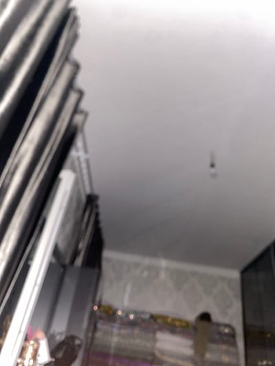 A close-up, blurry, high-angle shot captures a portion of a room. On the left, dark, vertical stripes suggest curtains or blinds. To the right, the ceiling is visible, with a small, out-of-focus light fixture hanging down. In the lower portion of the image, a blurry shelf or surface is visible with some indistinct objects on it, including what appear to be lights or reflective elements. The overall lighting is dim, contributing to the blurriness of the scene. The image does not contain any discernible information about the city of Боғистон, Tajikistan.