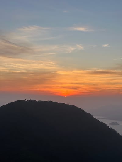 This media file captures a dramatic sunset over a mountainous landscape. The scene is divided between a vast sky and a silhouetted mountain range. The sky is painted with vibrant hues of orange and yellow, transitioning to softer pinks and blues towards the left. Wispy clouds are illuminated by the setting sun, creating streaks of light that stretch across the sky. The sun itself is a small, bright orange disc, partially obscured by the clouds and the mountain.

The foreground is dominated by the dark silhouette of a steep, jagged mountain. The outline of the mountain is sharp against the colorful sky, with some distant, fainter mountains visible in the hazy distance. The overall impression is one of natural beauty and tranquility, evoking a sense of awe at the grandeur of the sunset. The time of day is clearly sunset, given the intense colors in the sky. There is no visible text in the image. The location context provided is El Cacao, El Salvador.