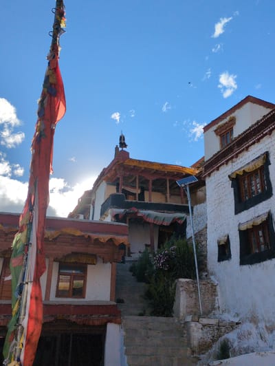 The image depicts a view of traditional Tibetan architecture in the city of Yulang, India, under a bright blue sky with scattered clouds. In the foreground, a tall pole with prayer flags is prominently displayed, the flags in vibrant colors of red, white, green, and yellow, adding a sense of cultural context. Behind the prayer flags, traditional buildings with whitewashed walls and intricate wooden window frames are visible. The architecture suggests a monastery or a traditional dwelling, characterized by layered rooftops and decorative elements. Sunlight streams from behind the buildings, creating a bright glare in the sky. In the middle ground, a solar panel is mounted on a pole, indicating a blend of traditional and modern infrastructure. The scene is devoid of any people, focusing solely on the structures and the environment. The overall atmosphere is serene and picturesque, highlighting the unique cultural and architectural heritage of the region.