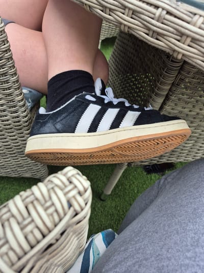 The image shows a close-up view of a person's legs and feet, wearing black socks and black Adidas sneakers with white stripes and laces. The person is seated on a wicker chair outdoors, with a green grassy surface visible beneath the chair and another wicker chair in the foreground. The lighting suggests it is daytime. There is no discernible connection to Carrick-On-Shannon, Ireland, as no landmarks or specific details are present to indicate this location.