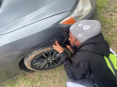 A man with dark skin, a beard, and a black hoodie is inspecting the front fender of a dark grey car. He is wearing a grey beanie with white flower patterns. The car has several scratches and scuff marks on its fender, especially near the wheel well. The tire is muddy, and the car appears to be parked on a grassy area with dirt. The man's hands are positioned as if he is feeling or examining the damaged area. In the background, a blurred grassy area suggests an outdoor setting. The lighting is bright, indicating it is daytime. The location context is Aiyegunle, Nigeria. There is no visible text in the image.