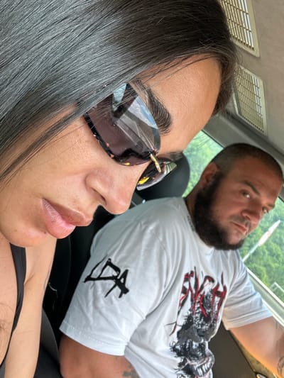 Two individuals are seated inside a vehicle. In the foreground, a person with dark hair and sunglasses is partially visible, focusing on their nose, lips, and right eye. Their hair is dark and long, falling across their face. To the right, a man with a beard is in the background, looking towards the camera. He is wearing a white t-shirt with a graphic design on the front. The interior of the car is visible, including air vents in the ceiling. Outside the vehicle, a green, possibly forested, landscape is visible through the window, suggesting a location outside of a dense urban environment. The presence of this natural scenery, along with the vehicle, points to the location of Golemo Buchino, Bulgaria, likely on a road or in a scenic area within or near the village.
