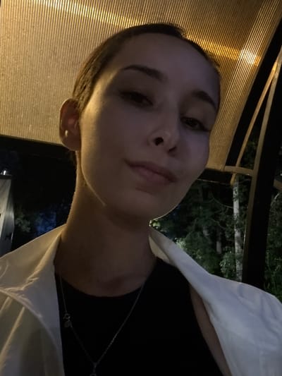 A close-up, low-angle selfie shows a young woman with her hair pulled back and wearing a black shirt and a white jacket. She is indoors, possibly on a patio or under a covered structure, with a slatted ceiling illuminated from above. The background is dark, with trees and foliage visible. The lighting suggests it is evening or nighttime. The woman is looking towards the camera with a neutral expression. The scene is located at Doma 1096 Km, Russia.