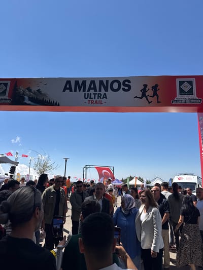 The image depicts an outdoor event, likely a running race, under a clear blue sky. A large banner with the text "AMANOŞ ULTRA - TRAIL -" and silhouettes of runners is prominently displayed. The banner also features logos and text related to "OSMANİYE BELEDİYESİ" (Osmaniye Municipality). In the foreground, a diverse crowd of people is gathered, some appearing to be participants or spectators. A few individuals are holding up mobile phones, possibly taking pictures or videos of the event. Tents and flags are visible in the background, suggesting a festive or organized gathering. The overall atmosphere appears to be one of community engagement and sporting activity in Osmaniye, Turkey.