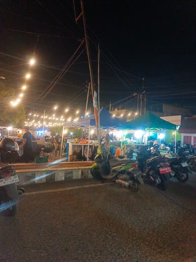 The image depicts a nighttime scene at an outdoor market in Luwoo, Indonesia. The market is illuminated by strings of overhead incandescent bulbs, casting a warm glow. Tents, likely for food stalls or vendors, are set up along a street. Several motorcycles are parked in the foreground and midground, some with their license plates visible. People are present within the market area, some appearing to be vendors and others customers. The overall atmosphere suggests a lively evening marketplace.