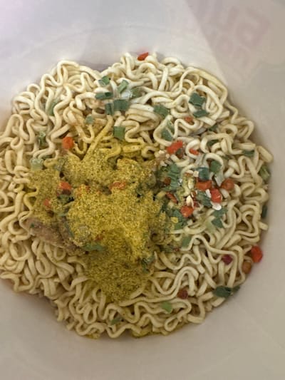 The image shows a close-up, overhead view of uncooked instant ramen noodles inside a white cup. The noodles are coiled and dry, with a pile of yellow seasoning powder and small pieces of dried vegetables (green onions, carrots, and possibly red chili flakes) on top. The background is a plain white, suggesting the interior of the cup or a simple surface. There are no people or any other objects visible. The image is a still life focused on the food product.