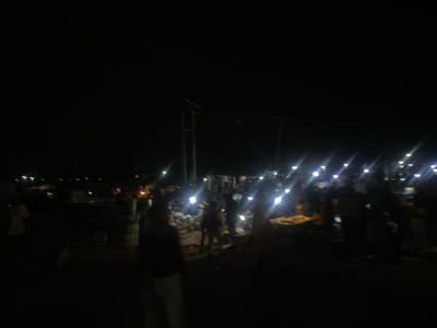 This media file depicts a nighttime scene in Sauka, Nigeria. The image is dark and blurry, suggesting it was taken in low light conditions, possibly with a handheld camera. The primary subjects are a group of people who appear to be gathered in an outdoor market or gathering place. There are numerous bright lights, likely from streetlights or market stalls, creating streaks and flares across the image due to the darkness and focus.
The setting seems to be an open area with some structures or stalls visible in the background, although details are obscured by the blurriness. Piles of objects, possibly goods for sale or discarded items, are scattered around. The activity taking place appears to be a lively evening market or social gathering, with people milling about and interacting.
The time of day is clearly night, indicated by the complete darkness of the sky and the artificial lighting. There are no clear weather cues, but the overall atmosphere suggests a warm evening, typical for the region. Location cues are limited to the provided context of Sauka, Nigeria.
No discernible text is visible in the image due to the low resolution and blurriness. The emotions conveyed are difficult to ascertain precisely, but the gathering of people under the lights suggests a sense of community and activity. The overall impression is one of a vibrant, albeit dimly lit, nocturnal scene in a Nigerian community.