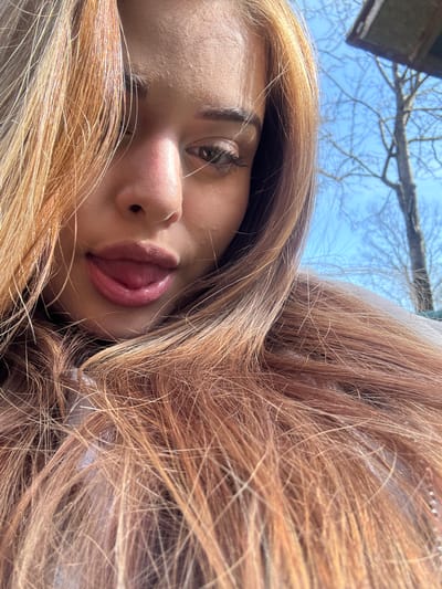 A young woman is lying down, possibly on a blanket or soft surface, with her head at an angle. Her long, wavy hair is spread around her, and her face is tilted towards the camera. She has fair skin, brown eyes, and full lips that are slightly parted, revealing the tip of her tongue. The background features a blue sky and bare tree branches. The location is not discernible from the image, and a determination of whether it is Gorno Trape, Bulgaria, cannot be made.