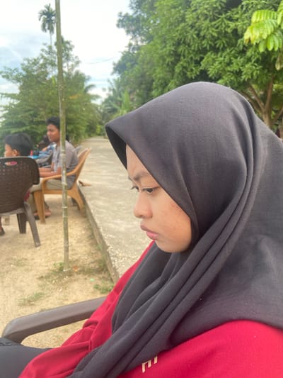 The image shows a young person wearing a black hijab and a red shirt, looking downwards. They have freckles on their face. In the background, there are trees and other people sitting on chairs, suggesting an outdoor gathering. One person in the background is wearing a blue and white checkered shirt. Another person, further in the background, is partially visible. There are several plastic chairs, some brown and some dark gray, scattered around. The ground appears to be dirt or sand, with some dry leaves. A concrete border runs along the background. The image is taken from a slightly elevated angle, looking down at the subjects. The scene appears to be set during the daytime, with bright lighting, and the foliage indicates a warm, possibly humid, climate. The overall atmosphere seems relaxed and casual. The location is identified as Air Molek, Indonesia. There is no visible text in the image.