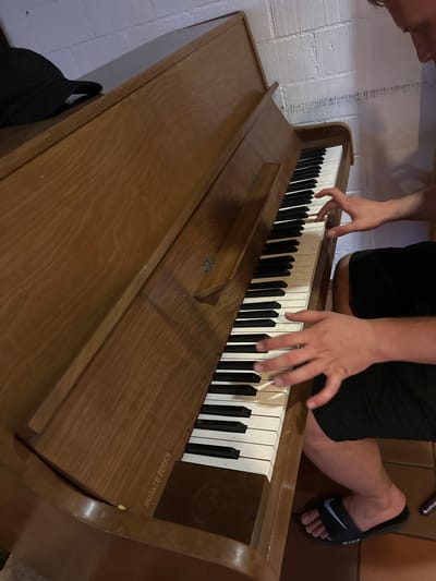 An overhead, slightly angled shot shows a person playing an upright piano in what appears to be a room with a brick wall in the background. The piano is made of wood and has white and black keys. The person's hands are visible on the keys, and they are wearing black shorts and black slide sandals with a Nike logo. A black baseball cap is on the piano lid to the left. The brand name "HUG & CO. ZÜRICH" is embossed on the wood below the keys. The overall lighting is dim, suggesting an indoor setting at night or in a low-light environment. There is no indication that this scene is located in the city of Wald (ZH), Switzerland.