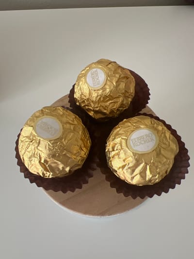 This media file features three Ferrero Rocher chocolates arranged on a light-colored surface, possibly a table or a platter. The chocolates are individually wrapped in crinkled gold foil, with a small white circular sticker on each, displaying the "FERRERO ROCHER" logo in brown lettering. Each chocolate is also nestled within a dark brown, fluted paper cup. The background is a plain, light gray, suggesting an indoor setting. The lighting appears to be natural, casting soft shadows and highlighting the metallic sheen of the chocolate wrappers. There are no people or animals visible in the image. The composition is a top-down view, with the chocolates forming a loose triangular arrangement. The overall impression is one of a simple, static display of confectionery.