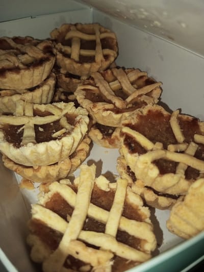 The image displays a quantity of small, round tarts with lattice tops, contained within an open, light-colored box. Each tart possesses a fluted, light-brown pastry crust holding a dark brown, viscous filling, surmounted by a crisscross pattern of thin, light-colored pastry strips. The tarts are arranged irregularly, appearing stacked and spread throughout the box, which presents as white or off-white cardboard with a potential dark teal or green edge visible on the lower right. Illumination from an unseen source casts minor shadows among the tarts. No individuals, actions, or specific interactions are discernible. The scene is located within a container holding confectionery items, reportedly in the city of Nam Mat, Laos.
