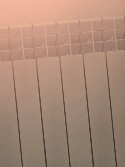 The image shows a close-up of a beige radiator, with vertical fins. The top of the radiator has a more intricate, segmented design, with curved edges visible. The background is a softly lit, pale pink or peach color, suggesting it's an indoor environment. The image is tightly cropped, focusing solely on the radiator's texture and form. There are no people or other objects visible in the frame. This image was taken in Avtury, Russia.