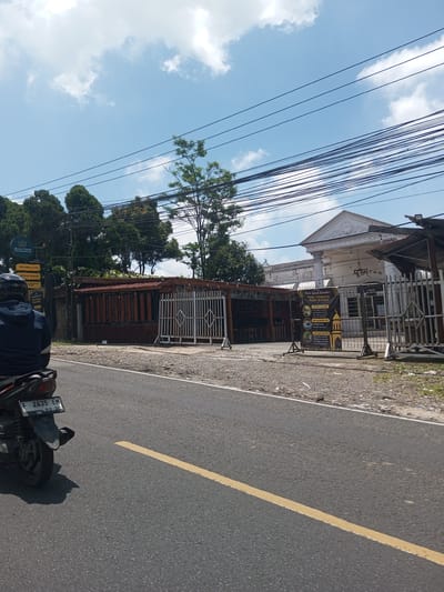 The image shows a street view with a motorcycle in the foreground. The motorcycle has a license plate "F 2635 EM 02-30." On the side of the road is a business complex identified as "Sultan" offering billiard & cafe, restaurant, karaoke, and Wi-Fi services. A sign advertises a Ramadan special menu. The architectural style of the building is distinct. Above the buildings are many wires and a partly cloudy, bright blue sky. The location is unspecified.