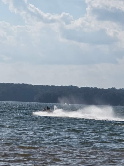 A jet ski is seen speeding across a body of water, creating a large spray behind it. The jet ski is ridden by a person who is silhouetted against the bright spray. In the background, a dense, tree-covered shoreline is visible, bordering the water. The sky above is filled with scattered clouds. The scene appears to be a recreational area, likely a lake or river within the city of Flowery Branch, United States of America, on a bright, possibly sunny day.