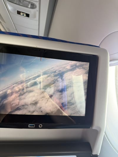 The image captures an interior view of an airplane cabin, focusing on an in-flight entertainment (IFE) screen attached to the back of a seat. The screen displays an aerial view, oriented sideways, showing a landscape of clouds, land, and what appears to be a body of water or coastline. Reflections are visible on the screen's surface, possibly from ambient cabin lighting or a window.

On the right bezel of the IFE screen, a circular headphone jack and a rectangular USB-A port are clearly visible. The screen is housed within a light gray plastic frame, which is part of the seat-back structure. A blue fabric strip with a subtle reddish pattern runs along the left side of the screen's frame, providing a visual accent.

To the left of the screen, parts of the airplane's interior ceiling are visible, including a light-colored panel with a dotted texture and a portion of an overhead compartment or light fixture with a reflective handle. Another seatback or partition is partially visible on the far right.

The scene depicts a passenger's experience during air travel, likely viewing content on the IFE system. No people are visible in the frame. The view on the screen suggests it is daytime. No discernible text is visible on the screen or surrounding cabin elements. The contextual location provided for the image is Parshino, Russia.